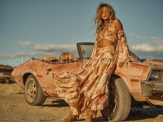 Boho styled woman in a long, patterned dress standing next to a rusted car in a desert setting